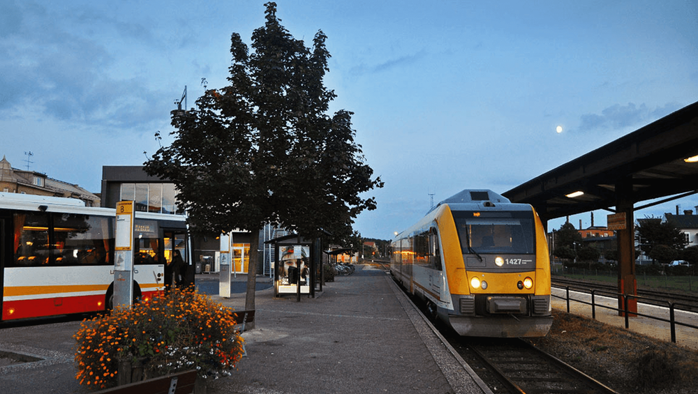 Dekorativ bild som visar Vetlanda station med ett tåg, en buss och grönska i form av buskar och träd.
